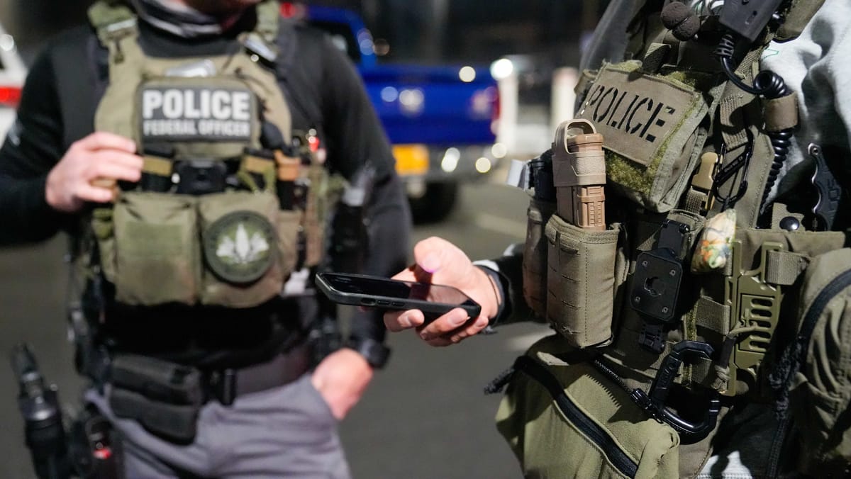 Two federal immigration police officers stand in tactical gear, with faces cropped from the photo, as one checks a phone.