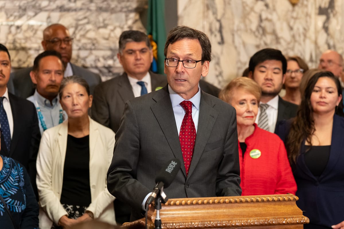 Washington governor Bob Ferguson stands at podium with legislators and Washingtonians behind him.