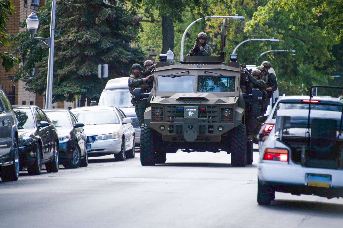An armored military vehicle with various ICE agents in tactical gear, drives down a wooded street lined with parked cars.
