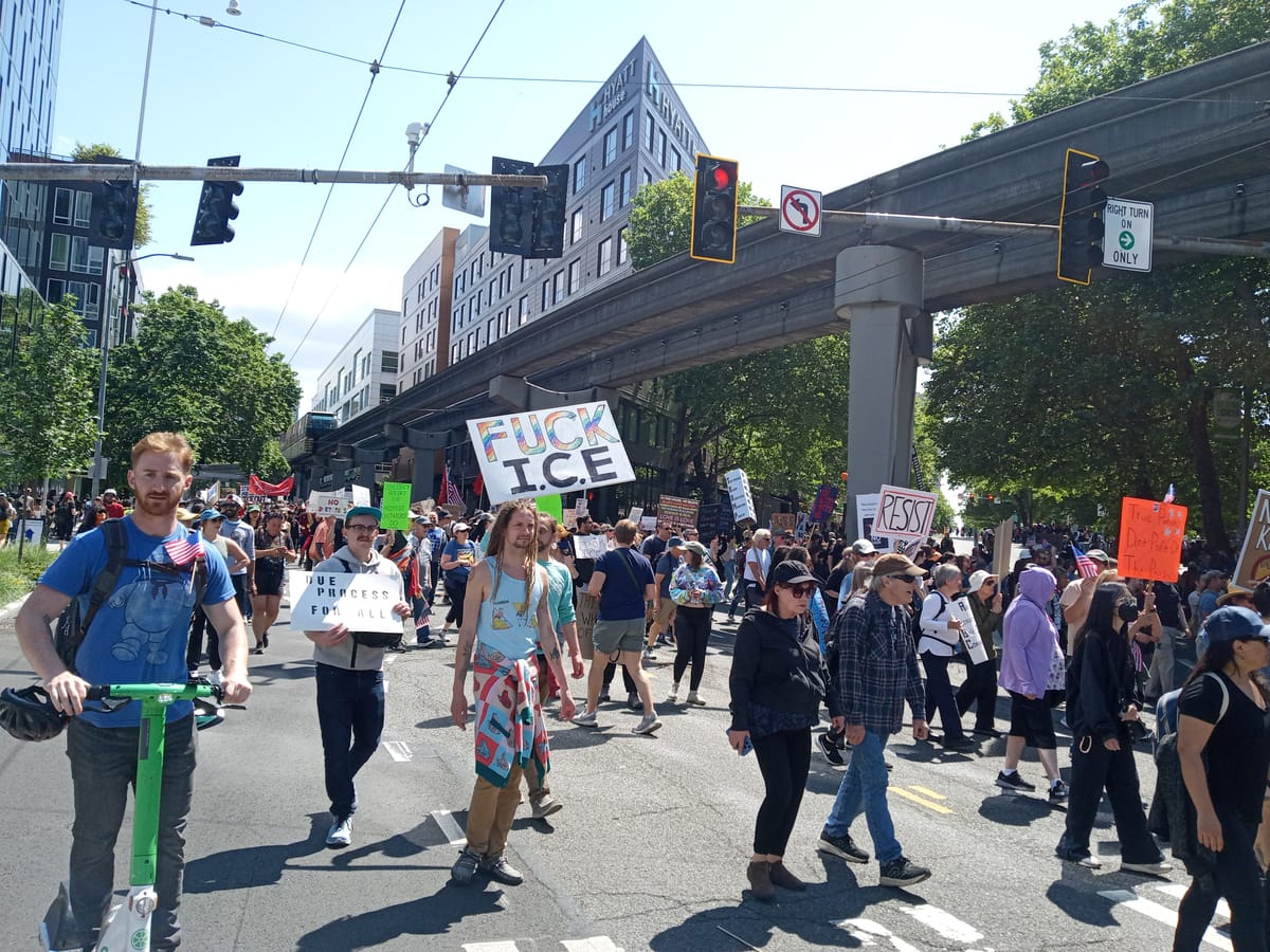 A crowd of protesters marches alongside the monorail with signs reading Fuck ICE and Resist.