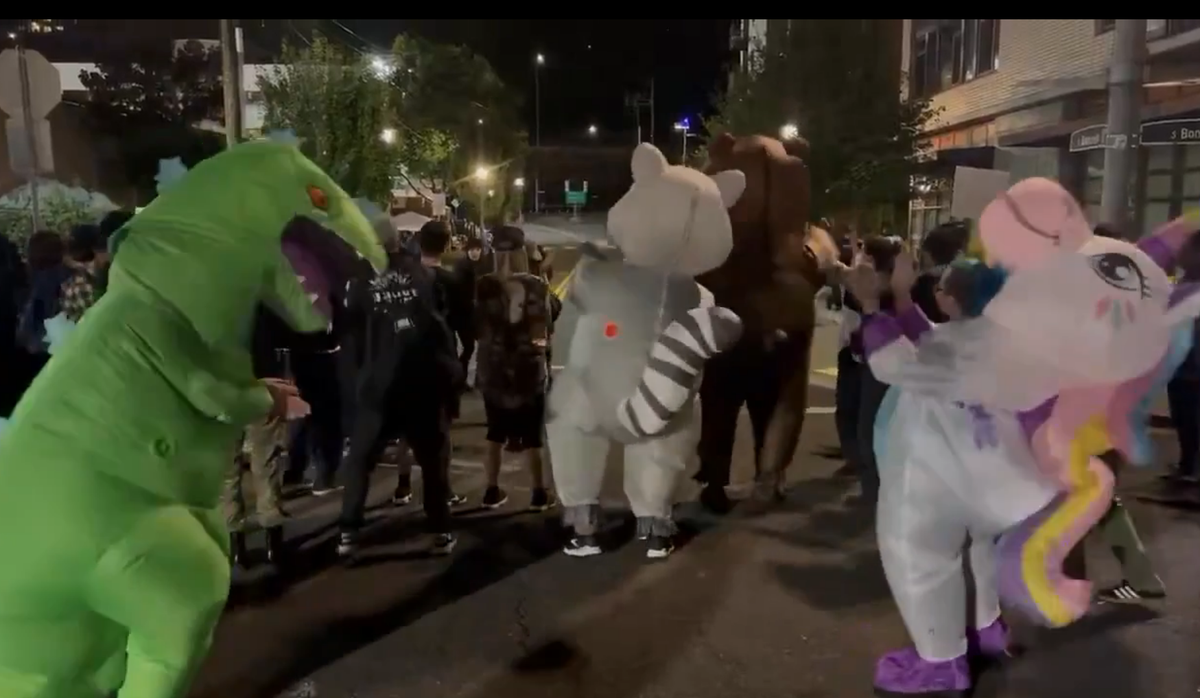 Protesters in inflatable Godzila, racoon, and unicorn suits dance in front of an ICE facility.