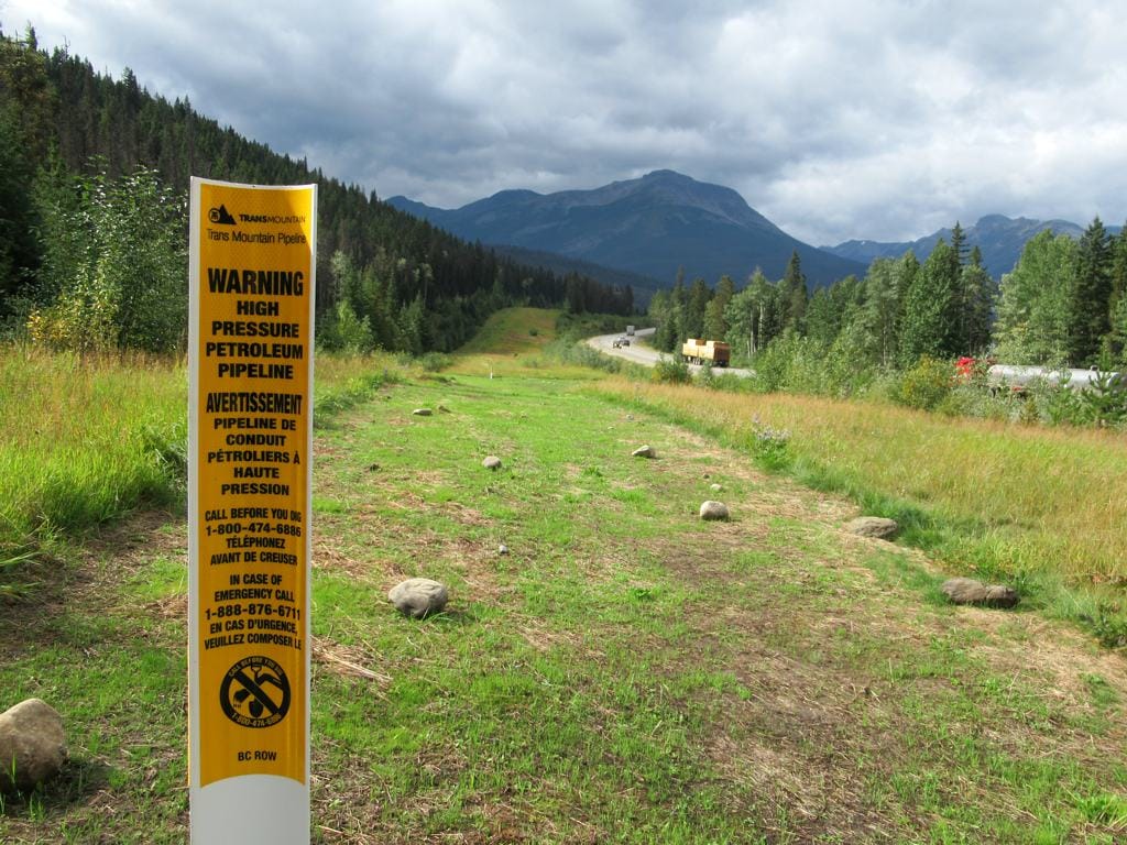 A sign reading Warning High Pressure Petroleum Pipeine in front of a clear pathway where the pipeline is buried, with mountains and a highway in the distance