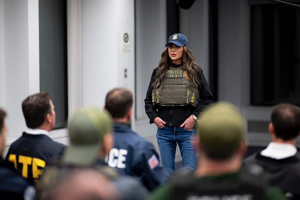 Kristi Noem, dressed in a bulletproof vest that reads Police ICE and a ballcap, speaks to a group of male federal officers.