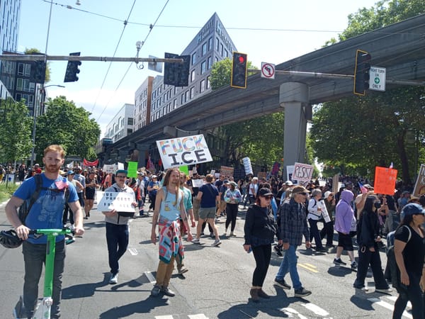 A crowd of protesters marches alongside the monorail with signs reading Fuck ICE and Resist.