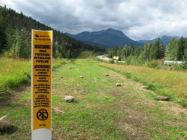 A sign reading Warning High Pressure Petroleum Pipeine in front of a clear pathway where the pipeline is buried, with mountains and a highway in the distance
