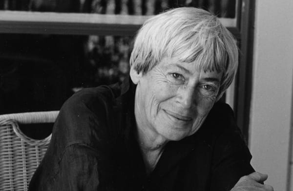 Writer Ursula LeGuin in a black and white image, sits in a wiker chair and smiles at the camera.